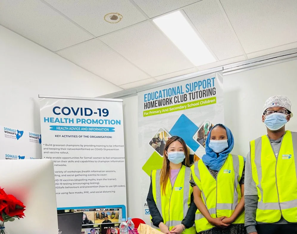 SWDA staff and volunteers in high-visibility vests beside COVID-19 health promotion and Homework Club tutoring banners